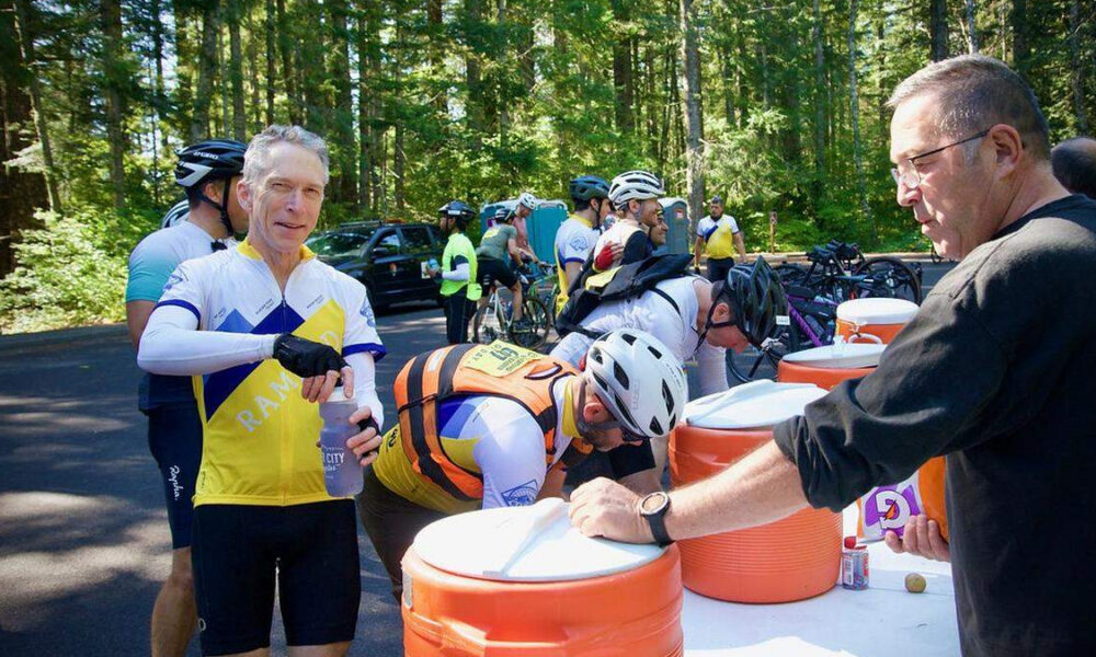 Cyclists at an outdoor water stop refilling bottles from large orange coolers during a group ride; trees in the background and volunteers assist.