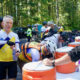 Cyclists at an outdoor water stop refilling bottles from large orange coolers during a group ride; trees in the background and volunteers assist.