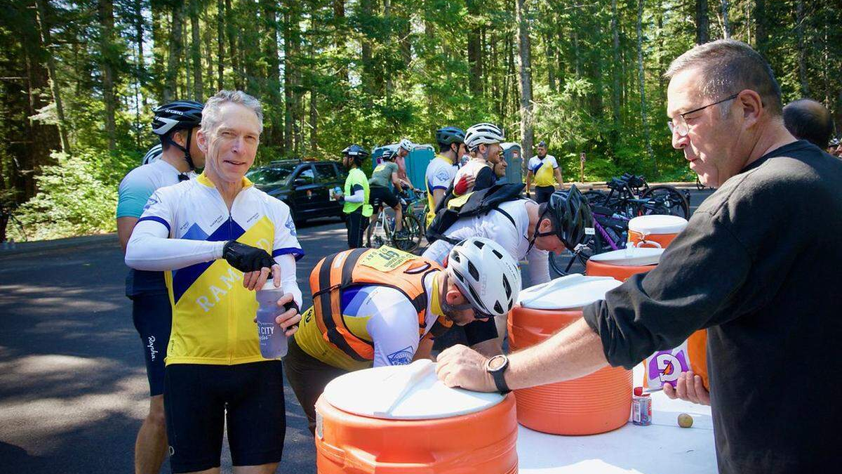 Cyclists at an outdoor water stop refilling bottles from large orange coolers during a group ride; trees in the background and volunteers assist.
