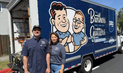 Two smiling gutter company workers pose in front of a blue Brothers Gutters truck with a cartoon logo; outdoor residential setting.