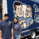 Two smiling gutter company workers pose in front of a blue Brothers Gutters truck with a cartoon logo; outdoor residential setting.