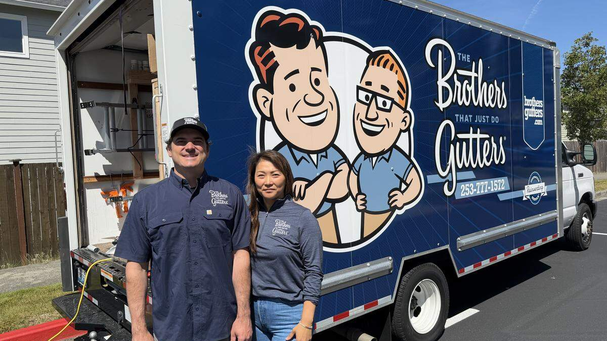Two smiling gutter company workers pose in front of a blue Brothers Gutters truck with a cartoon logo; outdoor residential setting.