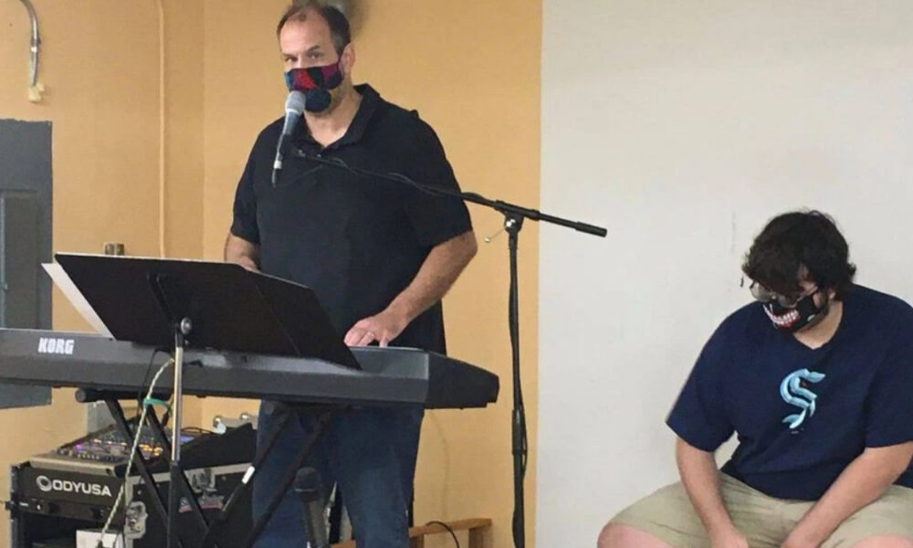 Two men wearing masks perform music: one at a KORG keyboard singing into a mic, the other seated nearby wearing a shark-mouth mask.
