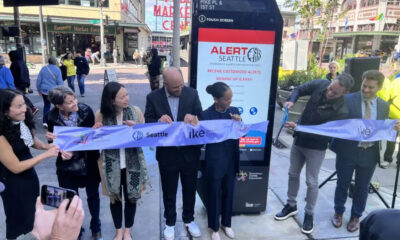 Group of six people cut a light-blue ribbon during a downtown Seattle ribbon-cutting ceremony, with a promotional display behind them.