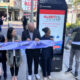 Group of six people cut a light-blue ribbon during a downtown Seattle ribbon-cutting ceremony, with a promotional display behind them.