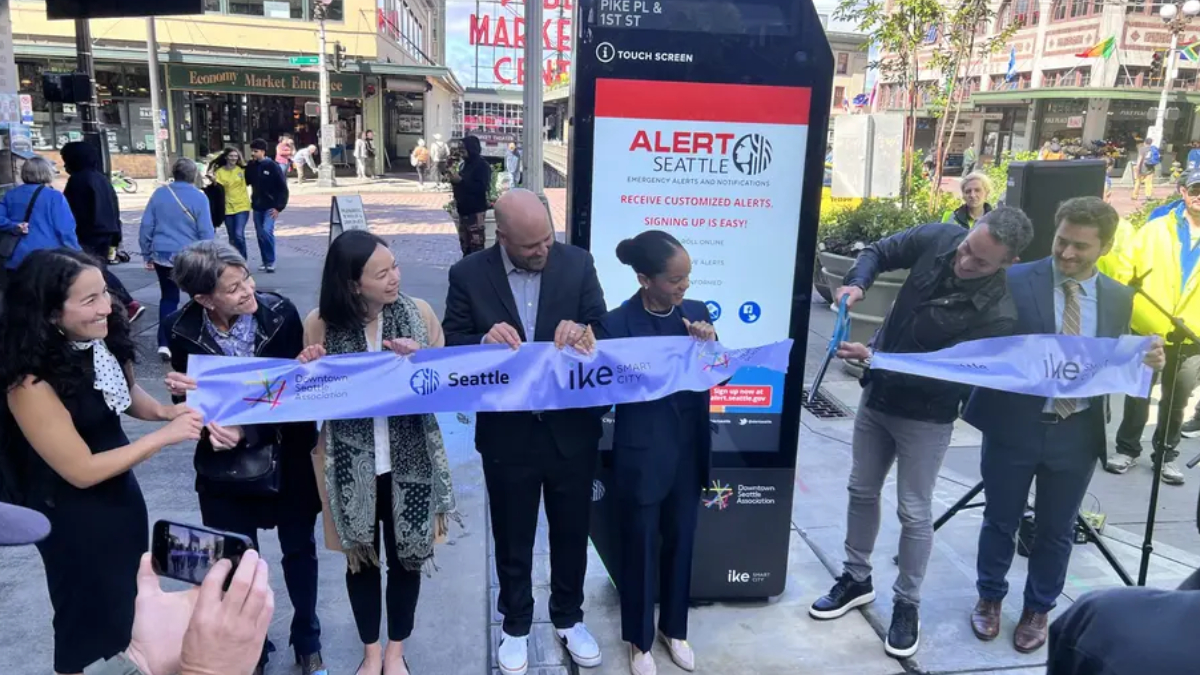 Group of six people cut a light-blue ribbon during a downtown Seattle ribbon-cutting ceremony, with a promotional display behind them.