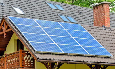 Blue solar panels installed on a sloped residential roof beside a brick chimney and wooden balcony railing