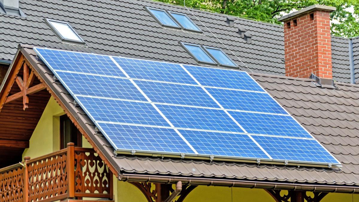 Blue solar panels installed on a sloped residential roof beside a brick chimney and wooden balcony railing