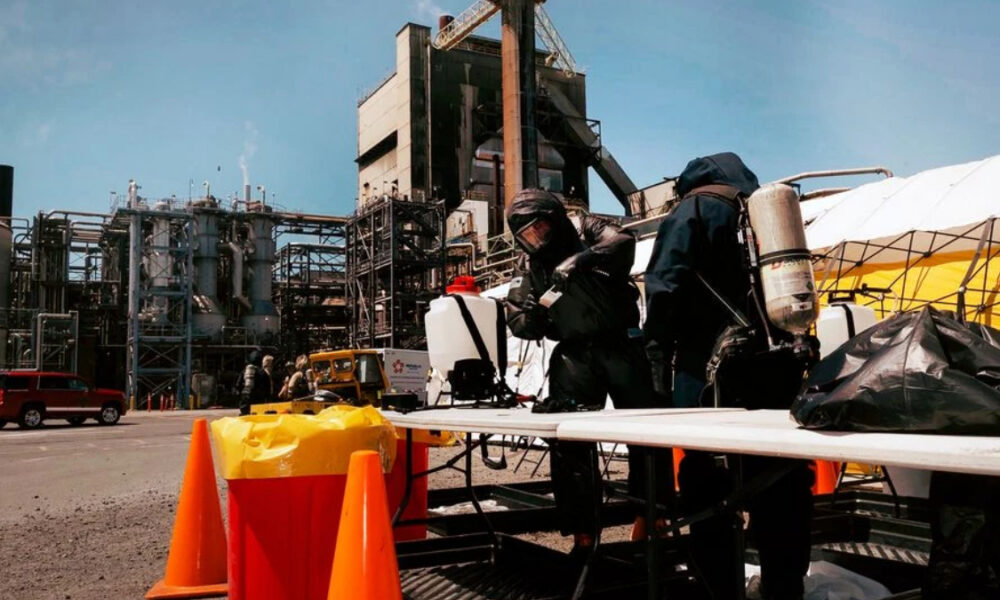 Two workers in full hazmat suits and breathing gear handle equipment at a chemical plant, with orange safety cones in the foreground and industrial structures in the background.