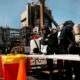 Two workers in full hazmat suits and breathing gear handle equipment at a chemical plant, with orange safety cones in the foreground and industrial structures in the background.
