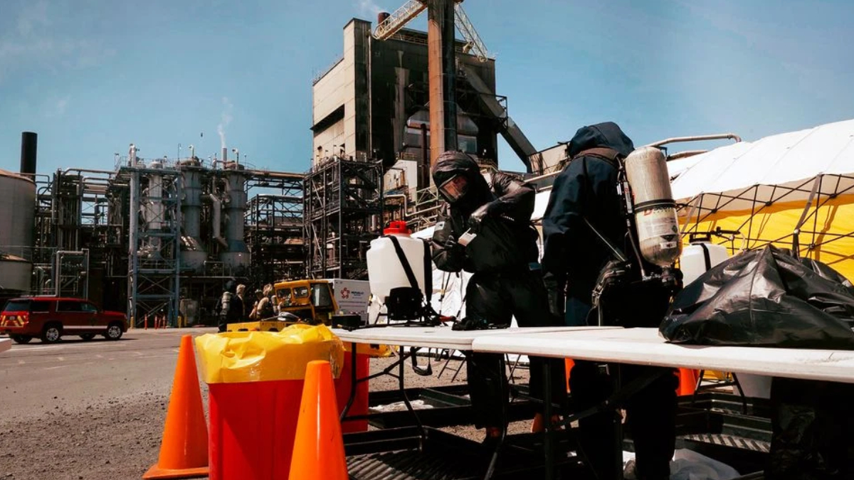Two workers in full hazmat suits and breathing gear handle equipment at a chemical plant, with orange safety cones in the foreground and industrial structures in the background.