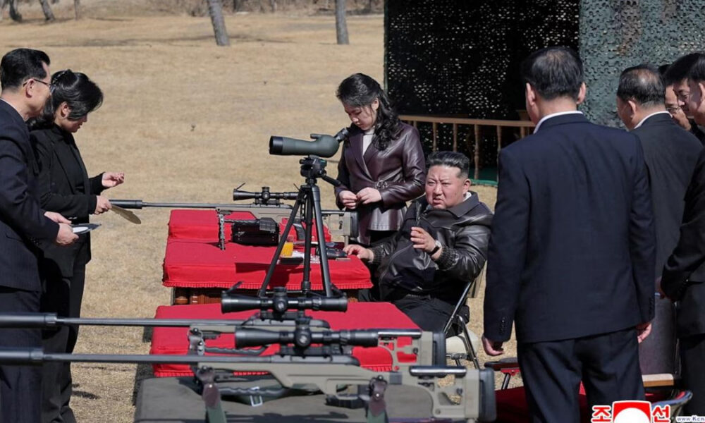 Man in a leather jacket sits at a table with a rifle and spotting scope while suited officials observe outdoors.