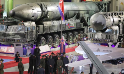 Display at a military exhibition featuring two large missile launchers and a rocket on a transport trailer, with visitors and officials nearby on a red carpeted floor.