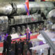 Display at a military exhibition featuring two large missile launchers and a rocket on a transport trailer, with visitors and officials nearby on a red carpeted floor.