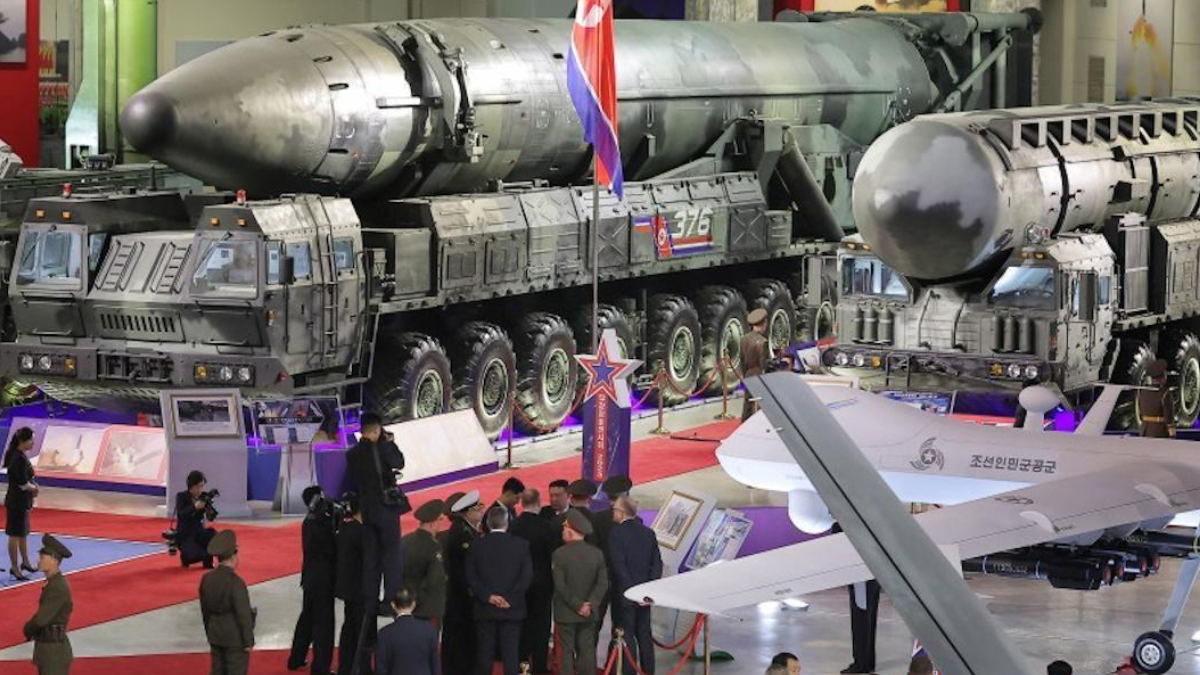 Display at a military exhibition featuring two large missile launchers and a rocket on a transport trailer, with visitors and officials nearby on a red carpeted floor.