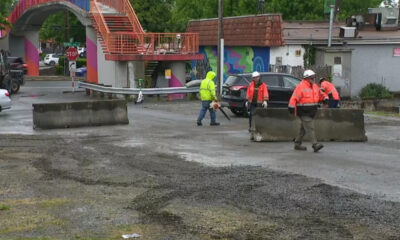 Construction workers in orange vests and hard hats directing traffic on a partially paved street with concrete barriers in the foreground.