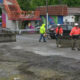 Construction workers in orange vests and hard hats directing traffic on a partially paved street with concrete barriers in the foreground.