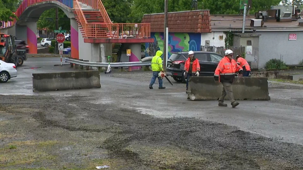 Construction workers in orange vests and hard hats directing traffic on a partially paved street with concrete barriers in the foreground.