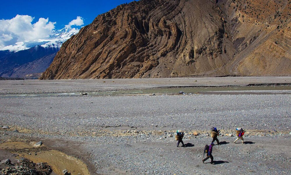 Four hikers with large backpacks trek across a gravelly desert plain, dwarfed by a steep brown mountain wall and distant snow-capped peaks under a blue sky.