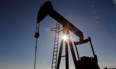 Silhouetted oil pumpjack against a clear blue sky with the sun peeking through its frame at sunset/sunrise.