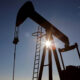 Silhouetted oil pumpjack against a clear blue sky with the sun peeking through its frame at sunset/sunrise.