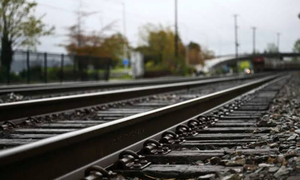 Perspective view of parallel railway tracks receding into the distance, with gravel and a blurred station area in the background.
