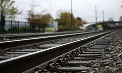 Perspective view of parallel railway tracks receding into the distance, with gravel and a blurred station area in the background.