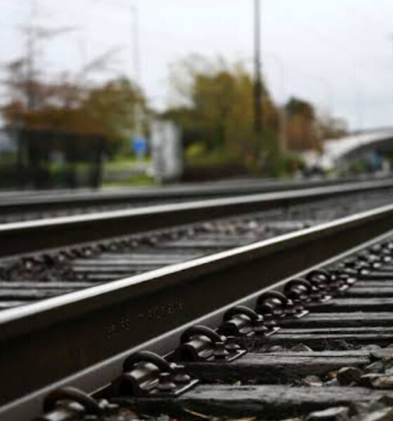 Perspective view of parallel railway tracks receding into the distance, with gravel and a blurred station area in the background.