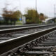 Perspective view of parallel railway tracks receding into the distance, with gravel and a blurred station area in the background.