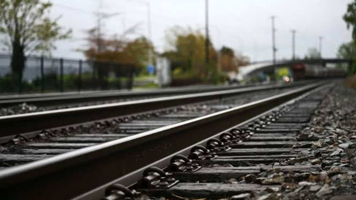 Perspective view of parallel railway tracks receding into the distance, with gravel and a blurred station area in the background.