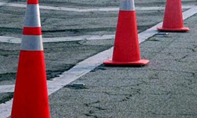Orange traffic cones create a barrier along a cracked asphalt road, marking a work area or detour on the street edge.