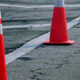 Orange traffic cones create a barrier along a cracked asphalt road, marking a work area or detour on the street edge.