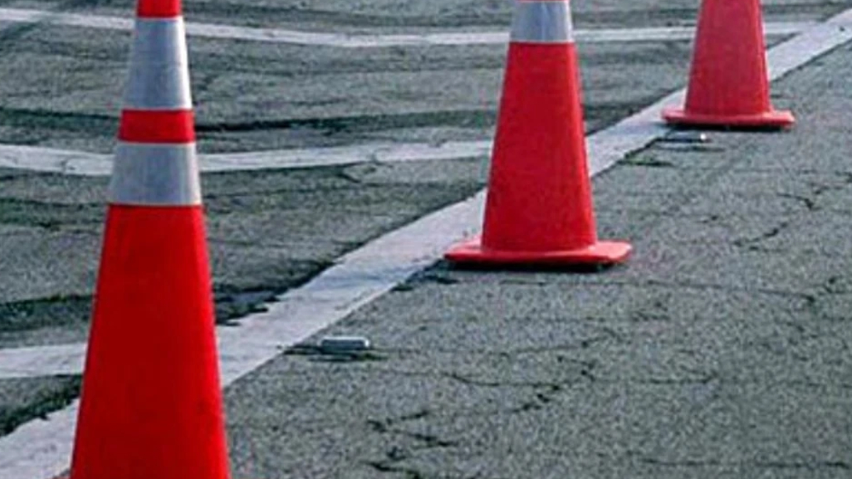 Orange traffic cones create a barrier along a cracked asphalt road, marking a work area or detour on the street edge.