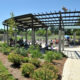 Public park pavilion with a lattice roof and concrete pillars, people sitting and walking on a sunny day in the background.