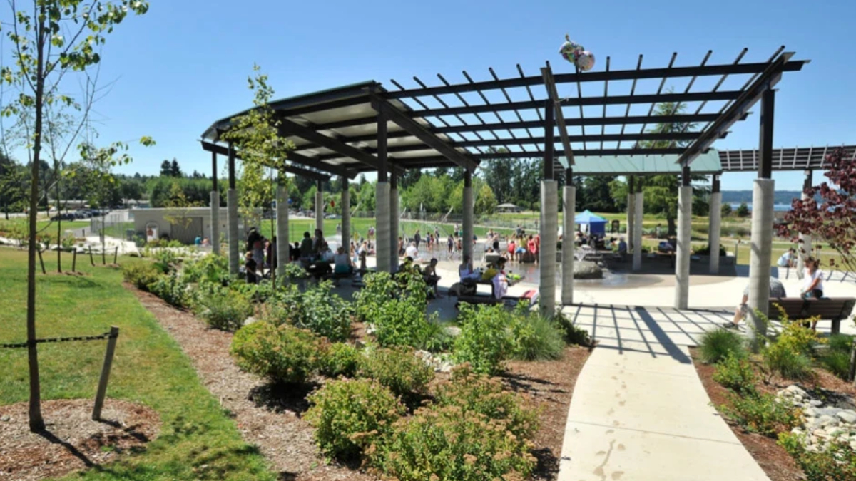 Public park pavilion with a lattice roof and concrete pillars, people sitting and walking on a sunny day in the background.