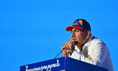 Man in a white shirt and blue cap with an orange Armenia map speaks into a microphone at a blue podium outdoors against a bright blue sky.
