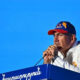 Man in a white shirt and blue cap with an orange Armenia map speaks into a microphone at a blue podium outdoors against a bright blue sky.