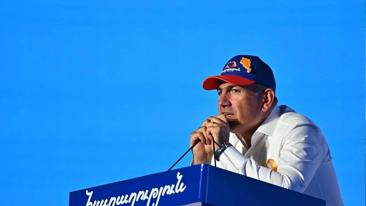 Man in a white shirt and blue cap with an orange Armenia map speaks into a microphone at a blue podium outdoors against a bright blue sky.