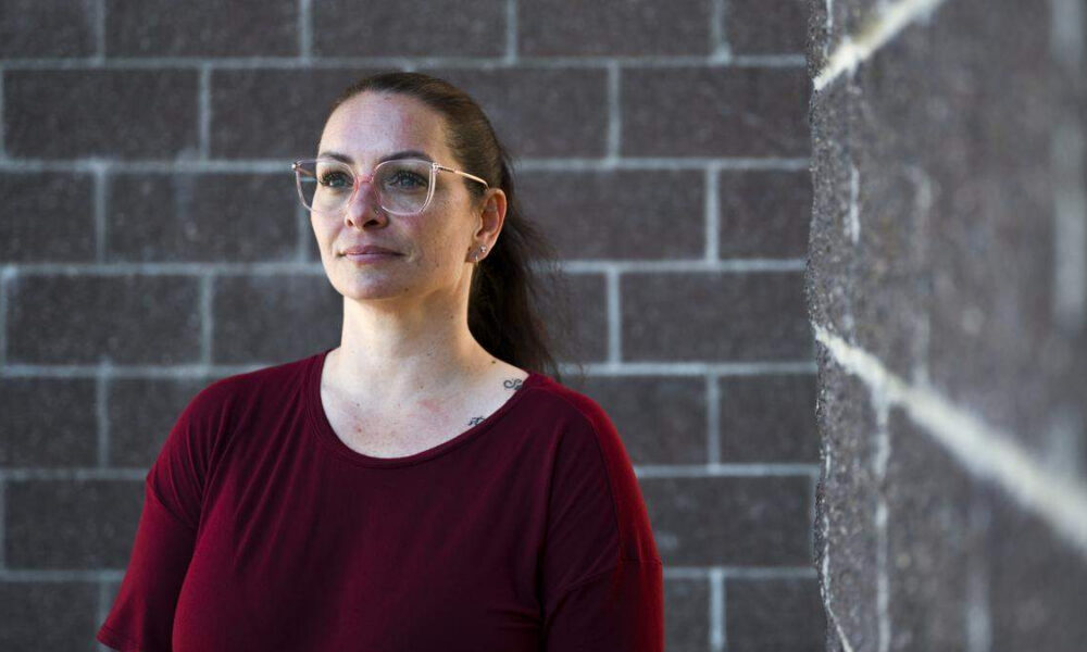 Woman with glasses and a burgundy shirt stands beside a gray brick wall, looking pensively into the distance.