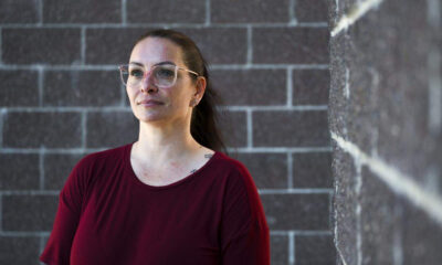 Woman with glasses and a burgundy shirt stands beside a gray brick wall, looking pensively into the distance.
