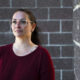 Woman with glasses and a burgundy shirt stands beside a gray brick wall, looking pensively into the distance.