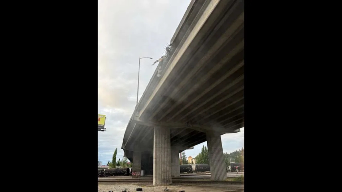 Damaged elevated highway with debris hanging from the edge; trucks and cars beneath as smoke rises nearby.