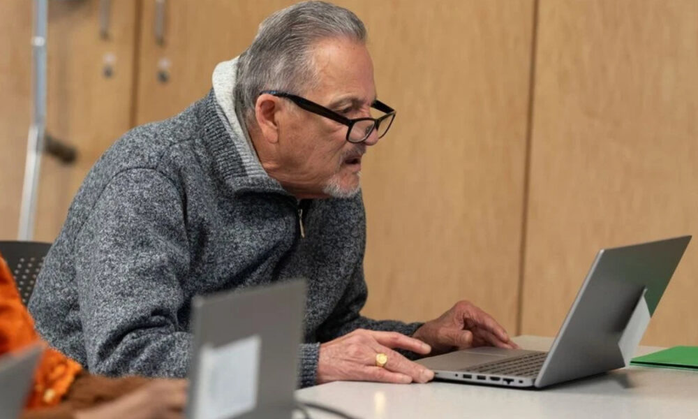 Older man with gray hair and glasses leans over a silver laptop on a table, focused on the screen in a meeting room setting.