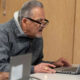 Older man with gray hair and glasses leans over a silver laptop on a table, focused on the screen in a meeting room setting.