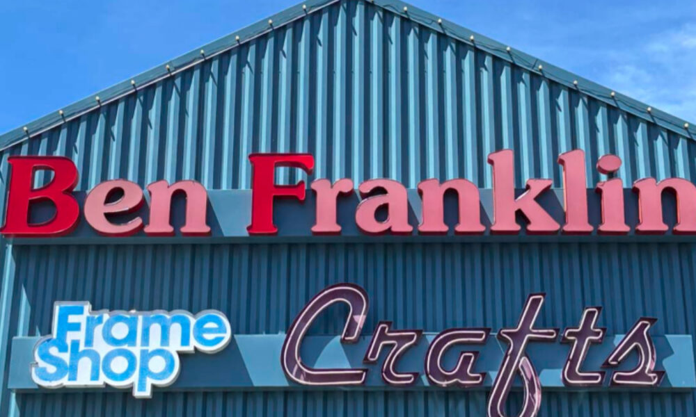 Storefront with a teal metal exterior and large red Ben Franklin sign above a blue and white 'Frame Shop' sign and cursive 'Crafts' lettering beneath.