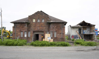 Abandoned brick building with boarded windows and overgrown shrubs; a yellow excavator sits to the left while a ruined structure stands to the right.