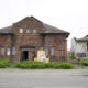 Abandoned brick building with boarded windows and overgrown shrubs; a yellow excavator sits to the left while a ruined structure stands to the right.