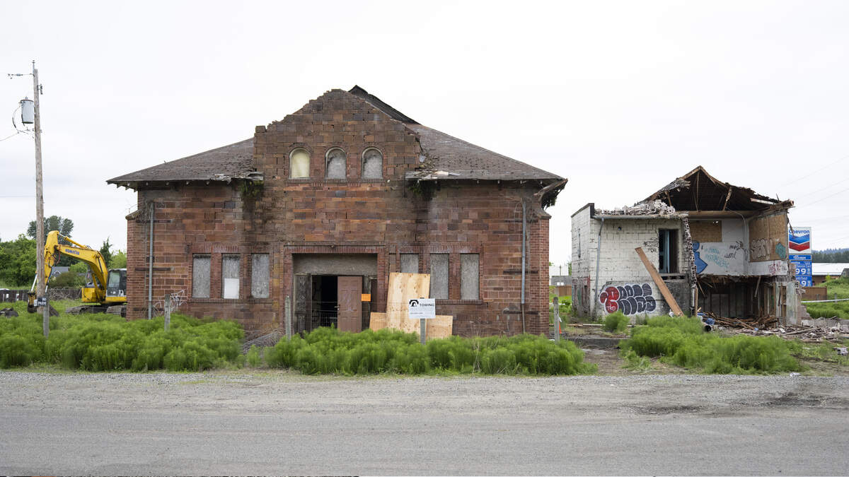 Abandoned brick building with boarded windows and overgrown shrubs; a yellow excavator sits to the left while a ruined structure stands to the right.
