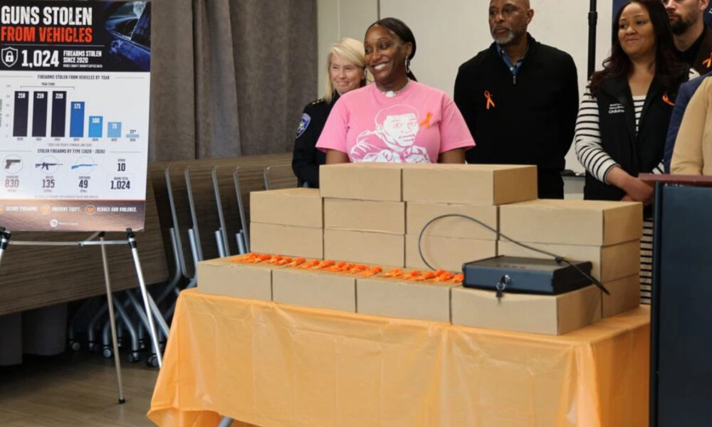 Group of people stand behind a table piled with brown cardboard boxes at a gun-violence awareness event, smiling.