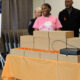 Group of people stand behind a table piled with brown cardboard boxes at a gun-violence awareness event, smiling.